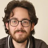 Ein junger Mann mit lockigem, dunklem Haar und einer Brille lächelt in die Kamera. Er trägt einen grauen Pullover mit einem besonderen Kragen. Der Hintergrund ist hell und schlicht.