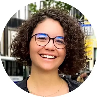 A smiling individual with curly hair and glasses is featured in a circular cropped image. They are outdoors, with a blurred city background suggesting urban surroundings. The person appears approachable and confident.