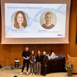 Eine Preisverleihung für den Annemarie Poustka Fellowship Award. Zwei Personen stehen in der Mitte der Bühne und halten einen Preis. Im Hintergrund stehen Kollegen und eine Präsentation mit Porträts der Preisträger ist sichtbar. Die Veranstaltung findet in einem Veranstaltungsraum statt.