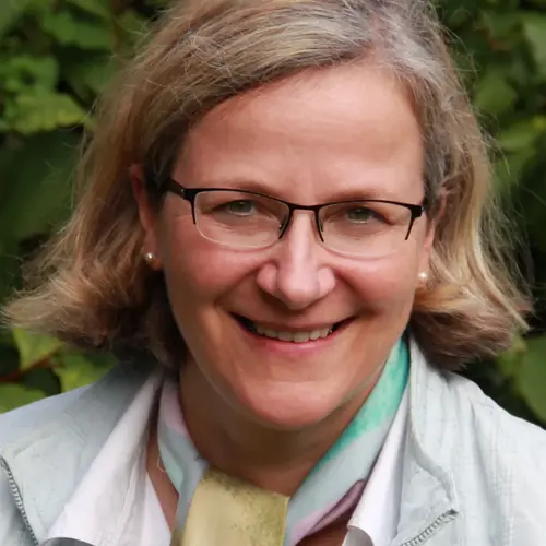 A woman with shoulder-length blonde hair smiles warmly at the camera. She wears glasses and is dressed in a light jacket over a white shirt with a colorful scarf. Lush green foliage serves as the background.