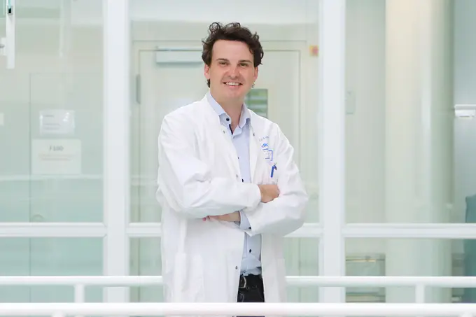 Jens Puschhof is wearing a white lab coat and standing with his arms crossed, smiling in a modern, bright environment. Behind him are large windows that offer a view of the hallway.