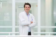 Jens Puschhof is wearing a white lab coat and standing with his arms crossed, smiling in a modern, bright environment. Behind him are large windows that offer a view of the hallway.