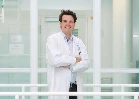 Jens Puschhof is wearing a white lab coat and standing with his arms crossed, smiling in a modern, bright environment. Behind him are large windows that offer a view of the hallway.
