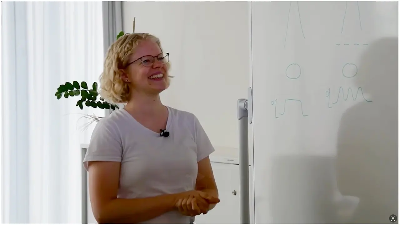 A smiling woman with curly hair and glasses stands beside a whiteboard. She is wearing a light shirt and appears engaged in a discussion. The whiteboard features simple drawings of human figures and lines. A plant is visible in the background.