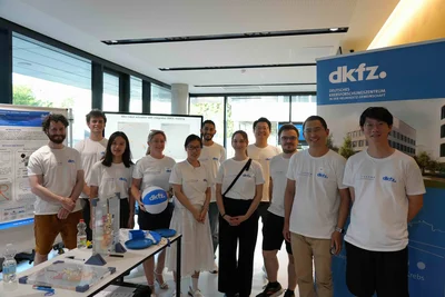 Eine Gruppe von zwölf Personen steht in einem modernen Raum. Alle tragen weiße T-Shirts mit dem Logo "dkfz". Im Hintergrund sind Poster und ein großes Banner zu sehen. Auf einem Tisch befinden sich verschiedene wissenschaftliche Materialien und Utensilien.