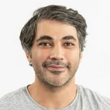 Ein lächelnder Mann mit dunklen, leicht gewellten Haaren und einem kurzen Bart trägt ein gestreiftes T-Shirt. Der Hintergrund ist neutral und hell.