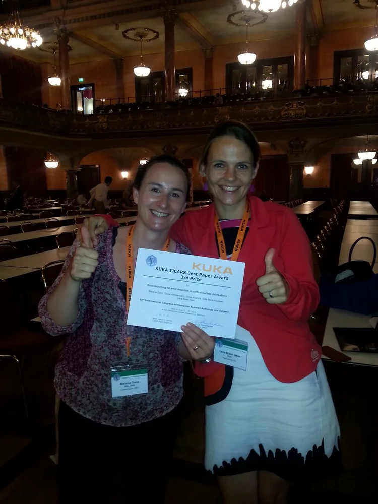 Two women stand smiling together in a large hall, each giving a thumbs-up. One holds a certificate titled "KUKA LUCAS Best Paper Award." The setting is formal, with rows of chairs and elegant lighting in the background, indicating a conference or award ceremony.