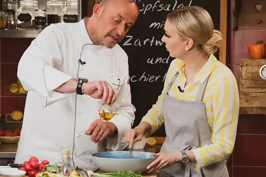Koch Thomas Kammeier steht neben TV-Moderatorin Susanne Klehn und füllt Öl in eine Salatschüssel.