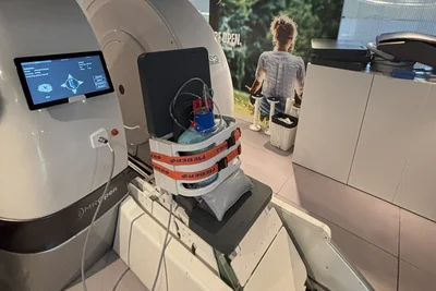 Eine MRT-Maschine mit einem Monitor, der Informationen anzeigt. Auf der Liege ist ein Patientensitz vorbereitet, der mit Riemen gesichert ist. Im Hintergrund ist ein Plakat mit einer Person, die im Freien sitzt.