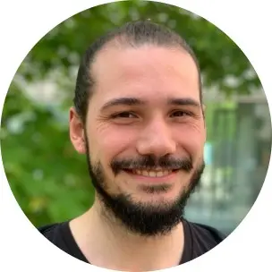 Portrait of a young man with a beard, smiling warmly. He has long dark hair pulled back and is wearing a black shirt. The background features soft greenery, suggesting an outdoor setting. This image represents Lukas Frank, an award recipient in a PhD program.