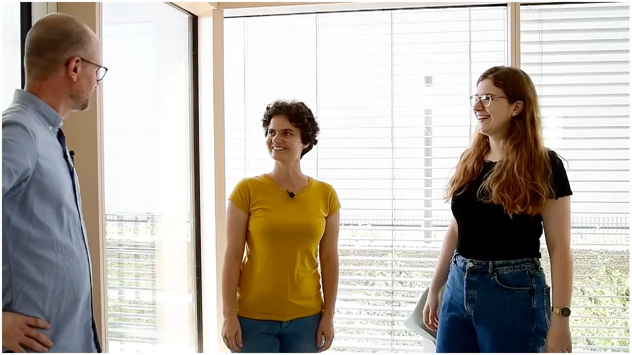 Three individuals stand in a well-lit room with large windows. One man, wearing glasses and a light blue shirt, faces two women. The woman on the left has short curly hair and wears a yellow shirt, while the woman on the right has long hair and wears a black shirt.
