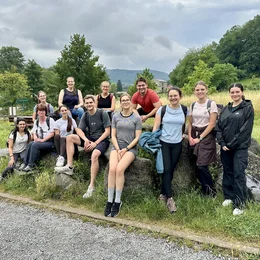 Eine Gruppe von 15 Personen steht und sitzt auf Steinen im Freien, umgeben von Bäumen und einer grünen Wiese. Es ist bewölkt, und die Stimmung wirkt entspannt und freundlich. Die Personen tragen Freizeitkleidung, einige scheinen zu lächeln oder mit ihren Freunden zu sprechen.