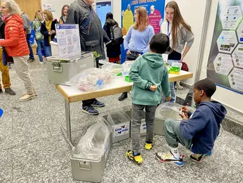 Zwei Kinder hocken auf dem Boden und sortieren Kunststoff in die passenden Metall-Autoklavenbehälter.