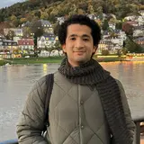 Ein Mann mit lockigem Haar steht an einem Flussufer, gekleidet in eine graue Jacke und einen gestrickten Schal. Im Hintergrund sind bunt gefärbte Bäume und mehrere Gebäude sichtbar. Die Szenerie ist während des Tages, mit leicht bewölktem Himmel.