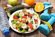 Ein Teller mit frischem Salat, der Salat, Tomaten, Gurken und Oliven enthält. Neben dem Teller liegen eine Gabel, eine Wasserflasche, ein Maßband und zwei blaue Hanteln. Im Hintergrund sind eine grüne Apfel und eine Orange sichtbar.