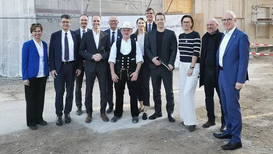 Eine Gruppe von zehn Personen steht auf einer Baustelle. Sie lachen und posieren gemeinsam. Im Mittelpunkt steht ein Mann in traditioneller Kleidung. Im Hintergrund sind Baustellenmaterialien und eine unvollendete Struktur sichtbar, was auf den Fortschritt eines Bauprojekts hinweist.