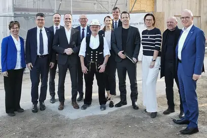 Eine Gruppe von zehn Personen steht auf einer Baustelle. Sie lachen und posieren gemeinsam. Im Mittelpunkt steht ein Mann in traditioneller Kleidung. Im Hintergrund sind Baustellenmaterialien und eine unvollendete Struktur sichtbar, was auf den Fortschritt eines Bauprojekts hinweist.