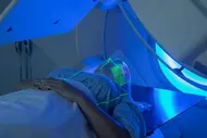 Ein Patient liegt auf einem Behandlungstisch in einer medizinischen Geräteeinrichtung. Grüne Laserlinienschnittmuster sind auf das Gesicht des Patienten projiziert, während eine blaue Beleuchtung um ihn herum leuchtet. Der Patient trägt ein Netz auf dem Kopf.