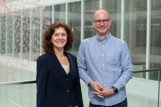 Two professionals stand smiling together in a modern indoor setting. They appear to be discussing or collaborating on topics related to particle therapy, reflecting the focus of the surrounding educational content on advancing training in this medical field.