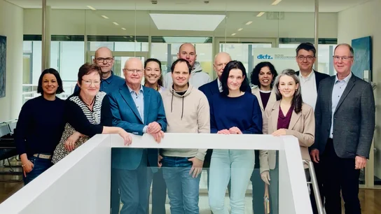 Die Mitglieder des Patientenbeirat Krebsforschung stehen gemeinsam mit dem DKFZ Vorstand und der Koordinatorin in einer Gruppe zusammen am Geländer einer Treppe und schauen in die Kamera.