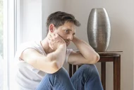 Ein nachdenklicher junger Mann sitzt auf dem Boden, die Hände hinter dem Kopf verschränkt. Er blickt nachdenklich zur Seite, während er an einem Tisch mit einer silbernen Vase neben einem Fenster sitzt.