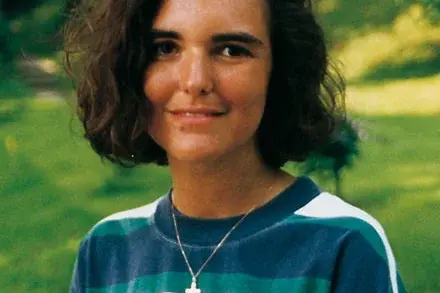 A young woman with curly hair stands outdoors, wearing a striped shirt with green, white, and blue colors. She looks directly at the camera and appears to be smiling softly, set against a blurred green background.