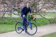 Ein junger Mann sitzt auf einem Fahrrad, das mit bunten Blumen geschmückt ist. Er lächelt und posiert in einem Park mit blühenden Bäumen im Hintergrund. Die Umgebung ist grün und einladend, ideal für einen Frühlingsausflug.