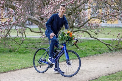Ein junger Mann sitzt auf einem Fahrrad, das mit bunten Blumen geschmückt ist. Er lächelt und posiert in einem Park mit blühenden Bäumen im Hintergrund. Die Umgebung ist grün und einladend, ideal für einen Frühlingsausflug.