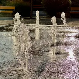 Ein Wasserspiel mit mehreren Fontänen, die in einem Park aufsteigen. Die Sonne reflektiert sich auf dem Wasser, während der Boden aus grauen Fliesen besteht. Um die Fontänen herum sind einige Bänke zu sehen, die zur Entspannung einladen.
