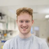 Ein junger Mann mit lockigem, rotblondem Haar lächelt freundlich in die Kamera. Er trägt einen grauen Pullover und steht in einem modernen Labor mit Regalen und Geräten im Hintergrund.