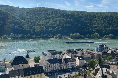 Eine Panoramaansicht eines kleinen Dorfes am Rhein, umgeben von grünen Hügeln. Im Vordergrund sind Häuser mit verschiedenen Dächern und Straßen zu sehen, während im Hintergrund der Fluss fließt und ein Schiff vorbeigefahren ist. Himmel blau mit leichten Wolken.