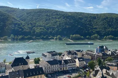 Eine Panoramaansicht eines kleinen Dorfes am Rhein, umgeben von grünen Hügeln. Im Vordergrund sind Häuser mit verschiedenen Dächern und Straßen zu sehen, während im Hintergrund der Fluss fließt und ein Schiff vorbeigefahren ist. Himmel blau mit leichten Wolken.