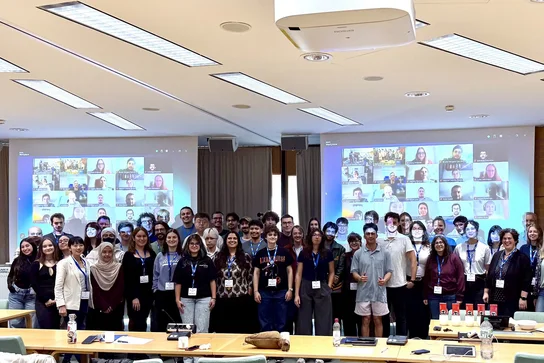 Eine Gruppe von mehr als 50 Personen steht in einem Konferenzraum und posiert für ein Gruppenfoto. Im Hintergrund sind mehrere Bildschirme sichtbar, die virtuelle Teilnehmer anzeigen. Alle Anwesenden tragen Namensschilder und sind vielfältig gekleidet.