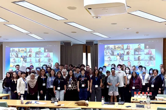 Eine Gruppe von mehr als 50 Personen steht in einem Konferenzraum und posiert für ein Gruppenfoto. Im Hintergrund sind mehrere Bildschirme sichtbar, die virtuelle Teilnehmer anzeigen. Alle Anwesenden tragen Namensschilder und sind vielfältig gekleidet.