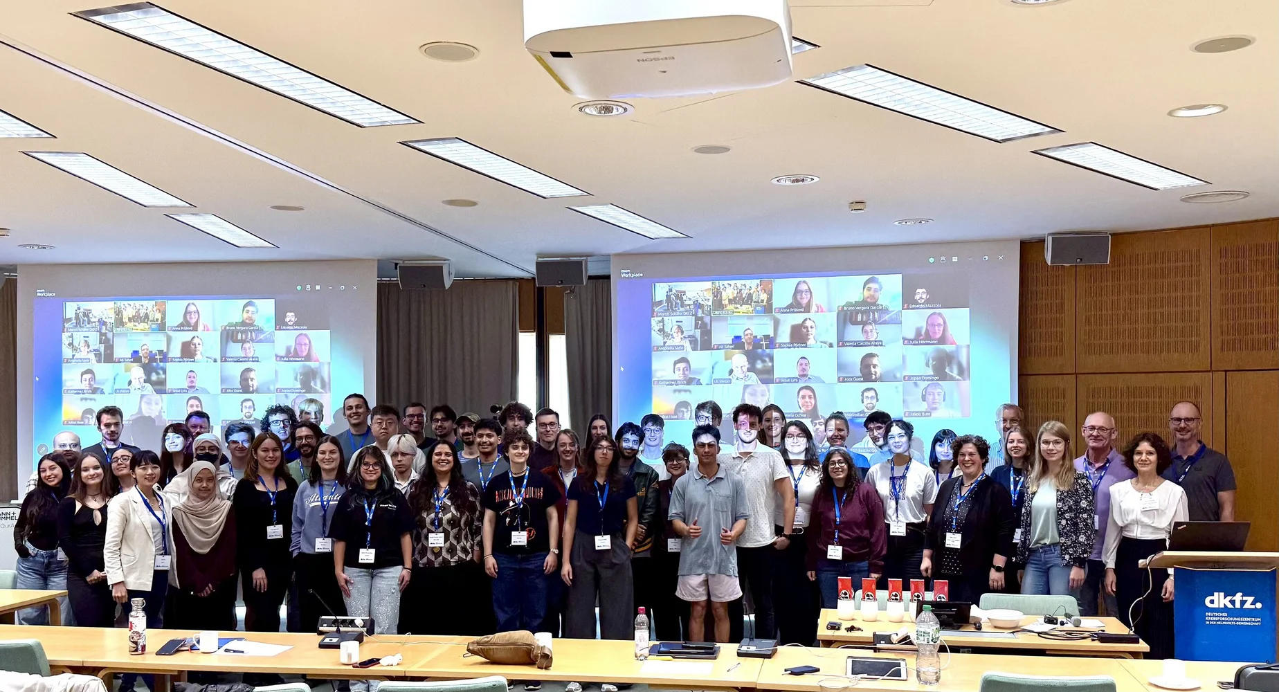 Eine Gruppe von mehr als 50 Personen steht in einem Konferenzraum und posiert für ein Gruppenfoto. Im Hintergrund sind mehrere Bildschirme sichtbar, die virtuelle Teilnehmer anzeigen. Alle Anwesenden tragen Namensschilder und sind vielfältig gekleidet.