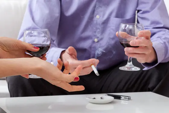 Zwei Personen sitzen an einem Tisch. Eine Hand reicht ein Glas Rotwein, während die andere Hand eine Zigarette hält. Auf dem Tisch befindet sich ein Aschenbecher. Die Stimmung wirkt entspannt und gesellig.