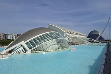 Moderne Architektur der Stadt der Künste und Wissenschaften in Valencia, Spanien. Futuristische Gebäude mit geschwungenen Formen und großen Glasfassaden, umgeben von blauen Wasserflächen und Rasen. Der Himmel ist klar und es gibt einige Wolken.