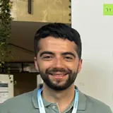 Ein junger Mann mit dunklem Haar und einem kurzen Bart lächelt in die Kamera. Er trägt ein graues Poloshirt und eine helle Umhängebandschleife. Hintergrund zeigt eine Wand mit Pflanzen und beschrifteten Papieren.