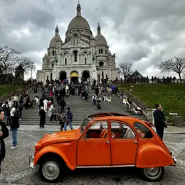 An orange vintage car is parked in the foreground, while a large crowd gathers at the steps of the Basilica of Sacré-Cœur in Paris. The basilica features stunning architectural details against a cloudy sky. People are mingling and sitting on the grass nearby.
