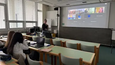 Eine Lehrerin steht vor einer Klasse und spricht. Auf einem großen Bildschirm sind online Teilnehmer zu sehen. Im Vordergrund sitzen Schüler an Tischen mit Laptops und Material. Der Raum hat große Fenster und ist hell, mit einem informellen Lernambiente.