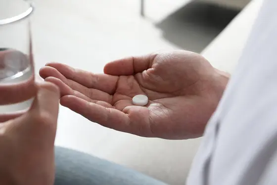 Eine Hand hält eine weiße Pille, während die andere Hand ein Glas mit Wasser ergreift. Die Szene deutet auf die Einnahme von Medikamenten hin.