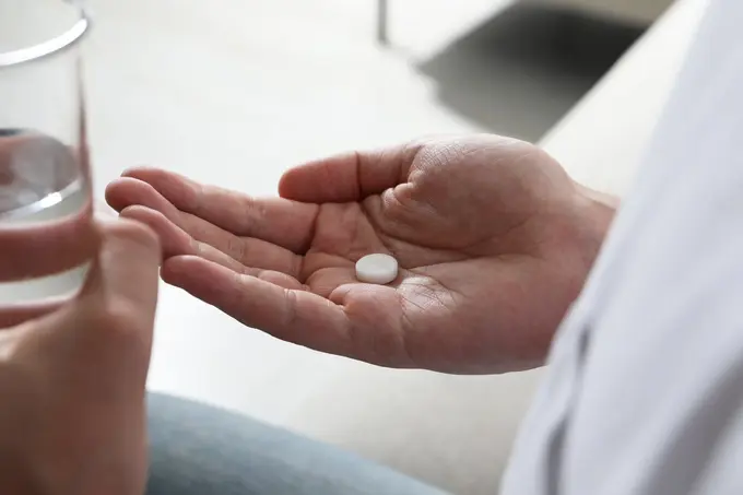 Eine Hand hält eine weiße Pille, während die andere Hand ein Glas mit Wasser ergreift. Die Szene deutet auf die Einnahme von Medikamenten hin.