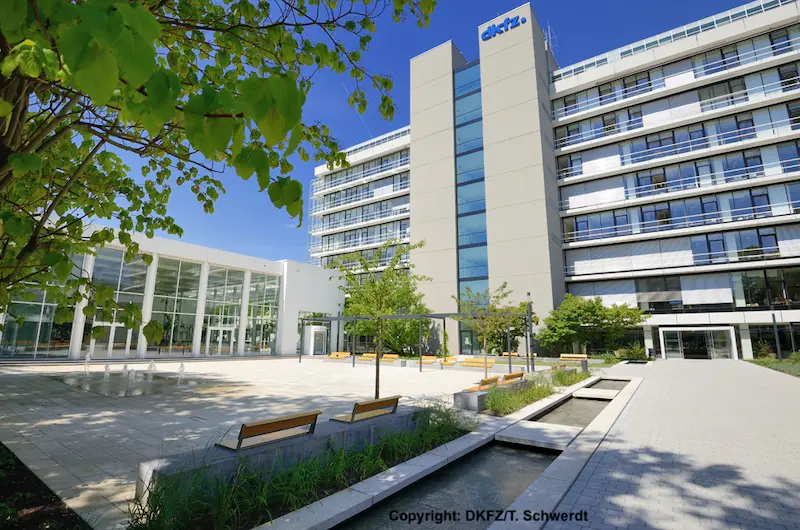 A modern building of the DKFZ (German Cancer Research Center) is depicted, featuring a glass entrance and surrounded by greenery. The image captures a clear blue sky and a paved area with benches, creating a welcoming atmosphere for visitors.