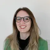 Ein Porträt einer jungen Frau mit langen, hellem Haar, die eine grüne Jacke und ein schwarzes Oberteil trägt. Sie lächelt freundlich in die Kamera und trägt eine Brille mit einem schwarzen Rahmen. Der Hintergrund ist neutral und hell.