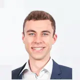 Ein junger Mann mit kurzen braunen Haaren und einem freundlichen Lächeln trägt einen blauen Anzug und ein weißes Hemd. Der Hintergrund ist hell und schlicht.