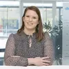 A smiling woman with long hair stands with her arms crossed, wearing a patterned blouse. She is positioned in a modern office environment with glass walls and plants in the background, conveying a professional and approachable demeanor.