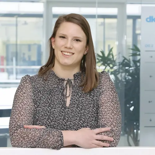 A smiling woman with long hair stands with her arms crossed, wearing a patterned blouse. She is positioned in a modern office environment with glass walls and plants in the background, conveying a professional and approachable demeanor.