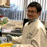 A smiling laboratory technician wearing a white lab coat and gloves measures a sample with a pipette. He stands at a lab bench with various scientific equipment and containers visible around him, emphasizing a professional and diligent work environment.