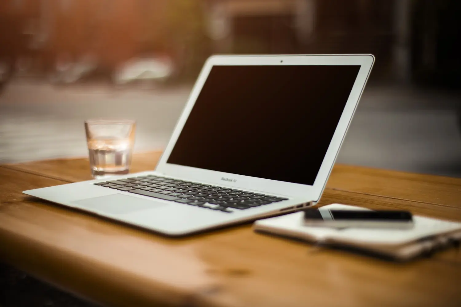 A sleek laptop sits open on a wooden table, with a notebook and smartphone nearby. A glass of water is placed next to it, creating a workspace vibe. The background is softly blurred, suggesting an outdoor setting. This image represents a modern, connected work environment.