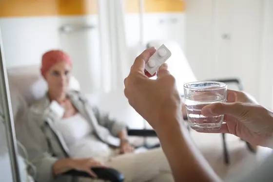 Ein Patient sitzt in einem Sessel, während jemand ihm ein Glas Wasser und Tabletten anbietet. Der Patient trägt ein Kopftuch und schaut aufmerksam auf das Angebot. Die Szene vermittelt eine Atmosphäre der Fürsorge und Unterstützung während einer Behandlung.